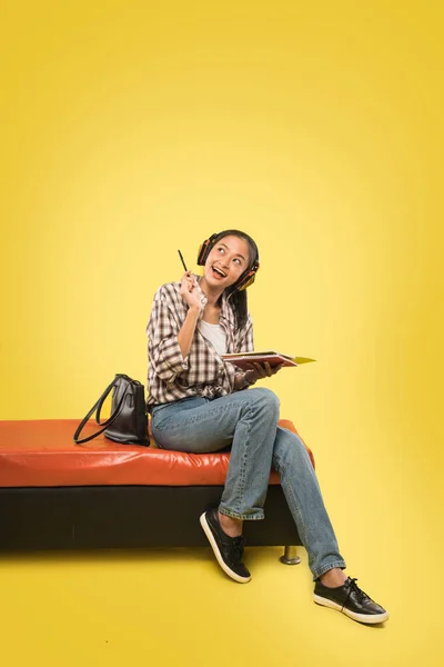 Hermosa chica sonriente con auriculares sosteniendo pluma y libro mientras mira hacia arriba con copyspace —  Fotos de Stock