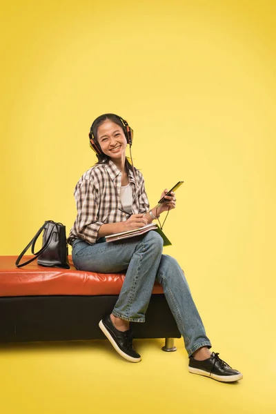 Χαριτωμένο κορίτσι γελώντας με κάμερα φορώντας ακουστικά και γράφοντας χρησιμοποιώντας στυλό στο βιβλίο, ενώ κρατώντας το κινητό τηλέφωνο — Φωτογραφία Αρχείου