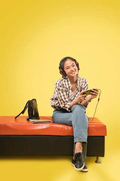 Ασιάτισσα με ακουστικά χρησιμοποιώντας smartphone, ενώ κάθεται με βιβλία και τσάντα — Φωτογραφία Αρχείου