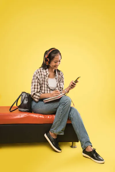 Mladá asijská dívka ve sluchátkách psaní v knize, zatímco pomocí mobilního telefonu sedí uvolněně — Stock fotografie