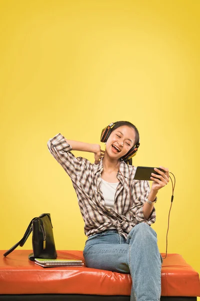 Gadis asia dengan headphone menggunakan smartphone sambil duduk dengan buku dan tas — Stok Foto