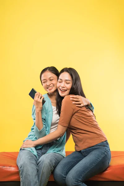 Close up of two young asian women laughing and hugging while joking sitting and one holding a mobile phone — Stock Photo, Image