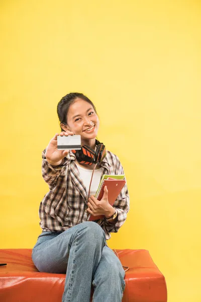 Sonriente joven mostrando tarjeta de crédito mientras sostiene libro y sentado —  Fotos de Stock