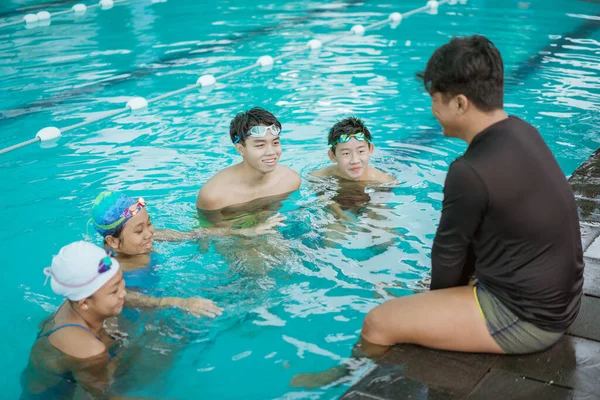 Instruktor v plaveckém tričku sedící u bazénu se čtyřmi teenagery v bazénu — Stock fotografie