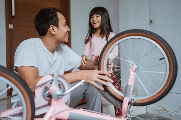 Córka śmieje się szczęśliwie, gdy jej ojciec naprawia swój mały rower. — Zdjęcie stockowe