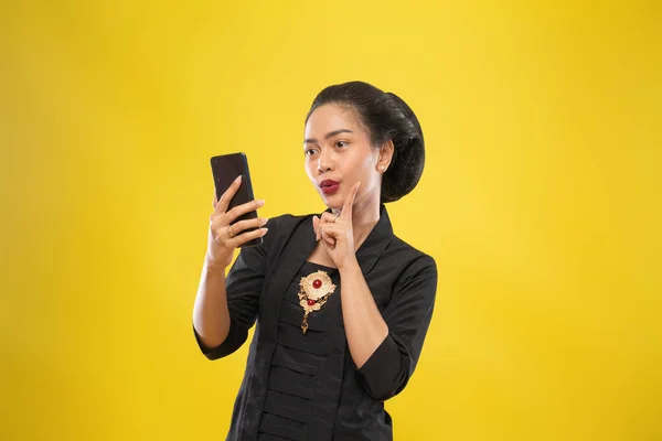 Javanese woman with traditional Kebaya dress thinking gesture holding smartphone — Stock Photo, Image