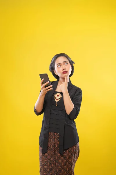 Donna giavanese con tradizionale Kebaya vestito pensiero gesto tenendo smartphone — Foto Stock