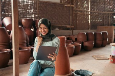 Tablet kullanarak çanak çömlek arasında oturan peçeli Asyalı kadın gülümsüyor.