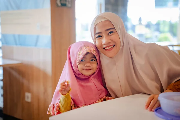 Muslim gadis muda sambil duduk di ruang makan dengan ibu tersenyum ke kamera — Stok Foto