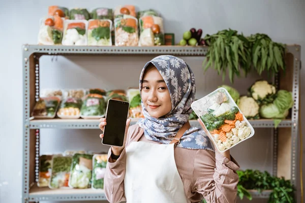 Vrouw groenteboer toont groenten en toont een telefoon scherm — Stockfoto
