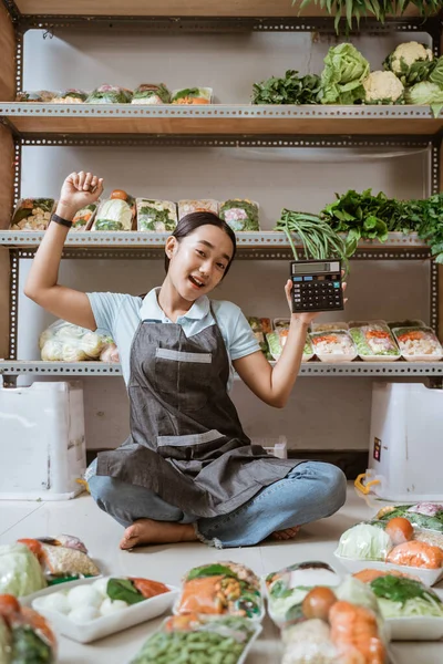Opgewonden veggie verkoper in schort zit op de vloer met behulp van rekenmachine — Stockfoto