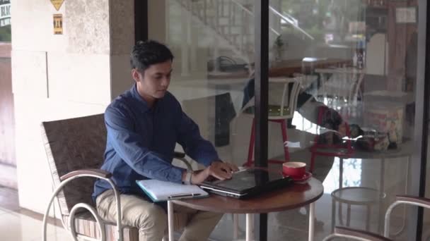 Aziatische man met zeggen hallo gebaar tijdens het werken met behulp van laptop — Stockvideo