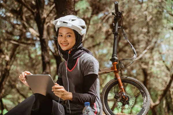 Mujer velada sentada descansando en bicicleta usando auriculares y usando tableta —  Fotos de Stock