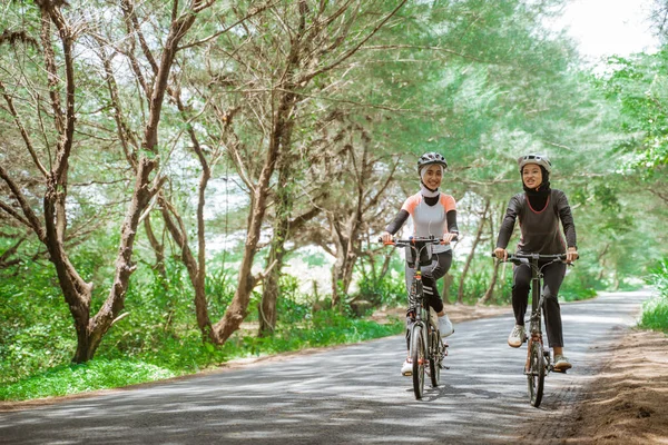 Two asian muslim women cycling on the road many trees — стоковое фото