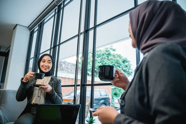 Two asian muslim woman talking while sitting on a couch enjoying coffee — стоковое фото