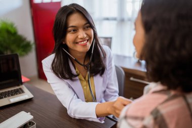 female doctor smiling while examining female patients chest using a stethoscope in clinic room
