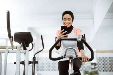 attractive muslim woman using mobile phone during exercising with static bike at the gym