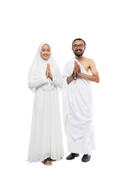 Husband and wife wearing white hajj ihram clothes with greeting gesture standing on isolated background