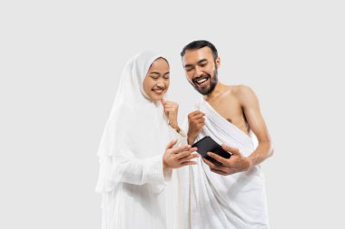 Excited couple wearing ihram clothes looking at video using smartphone on isolated background