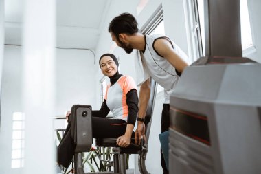 Personal trainer helping woman with hijab working with leg weight machine at the gym