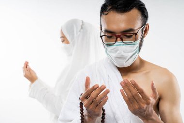 Man in ihram clothes and mask praying with prayer beads with a woman praying in the back on white background