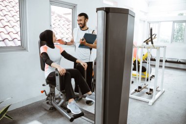 Personal trainer helping woman with hijab working with leg weight machine at the gym