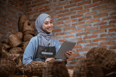 craftswoman wearing hijab smiling while using a tablet to sell water hyacinth handicrafts online at a village house