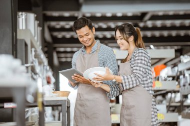 Man in apron using pad to check things with woman while holding plate in houseware store