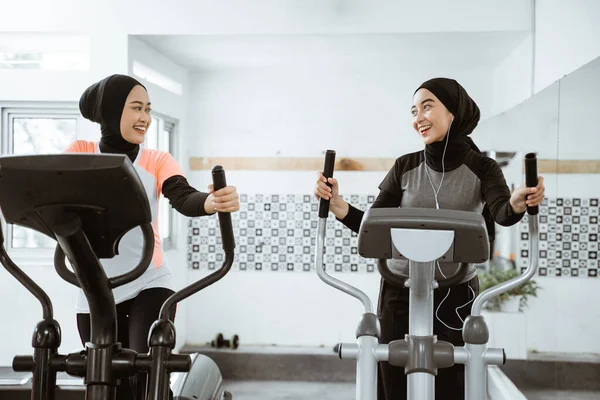 two beautiful woman with hijab at the gym exercising with friend on static elliptical cycle machine together