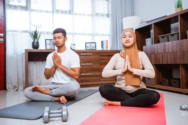 muslim beautiful woman and husband doing yoga at home together. couple exercising in livingroom