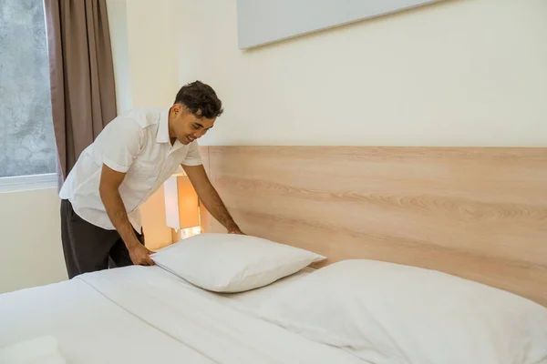 young male housekeeper tidying pillows on bed in hotel room