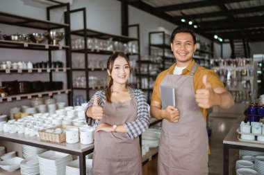 Two shop owners in apron holding digital tablet standing with thumbs up in houseware store
