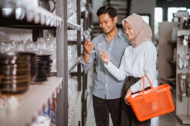 Couple of customers holding glasses while selecting glassware on shelf in houseware store