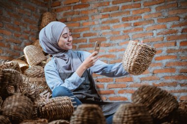 craftswoman in veil photographing using mobile phone handmade woven water hyacinth in brick house