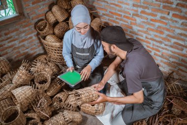 top view of craftswoman in veil and craftsman using tablet among woven water hyacinth crafts in brick house