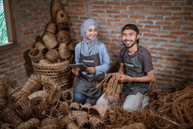 smiling woman in veil using tablet and smiling craftsman holding woven rope in brick house
