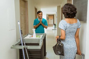 female hotel guest meets a janitor with a greeting gesture pushing a cart filled with cleaning supplies