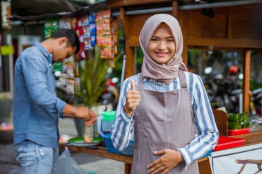 proud young muslim woman standing in front of her small traditional food shop and showing thumb up oke gesture clipart