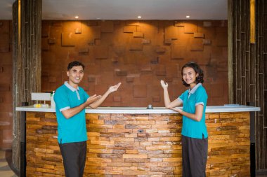 two employees wearing turquoise uniforms smiling with hand gestures presenting something at the reception desk