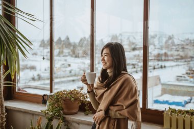 beautiful asian woman enjoy coffee morning standing next to a big window with beautiful view of snowy landscape in cappadocia