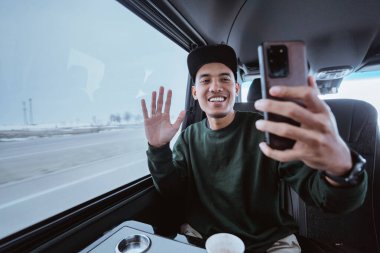 young asian male making a video call and selfie during his way out. man using mobile phone while travelling by a van