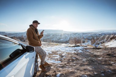 man using his mobile phone at the top of the hill while leaning against his car with beautiful view at the background