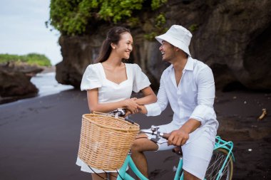happy asian young couple riding bicycles on black sandy beach
