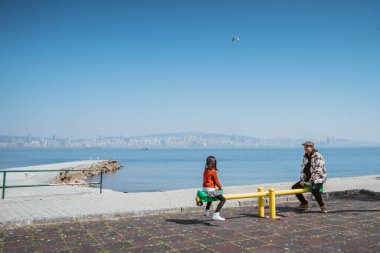 father and daughter playing seesaw together in playground with sea background