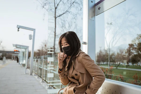 young woman coughing while sitting on train station wearing face mask