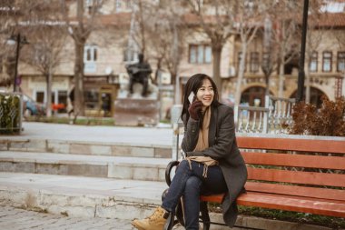 asian woman sitting on a bench in the park while using her mobile phone