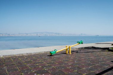 portrait of a seesaw in empty playground next to the sea with a city at the far back