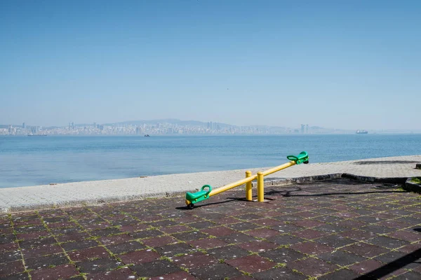 portrait of a seesaw in empty playground next to the sea with a city at the far back