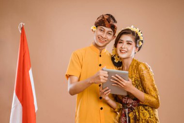 man and woman wearing balinese traditional clothing using tablet pc and smiling