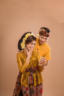 suprised balinese couple with kebaya while looking at her mobil phone screen over isolated background